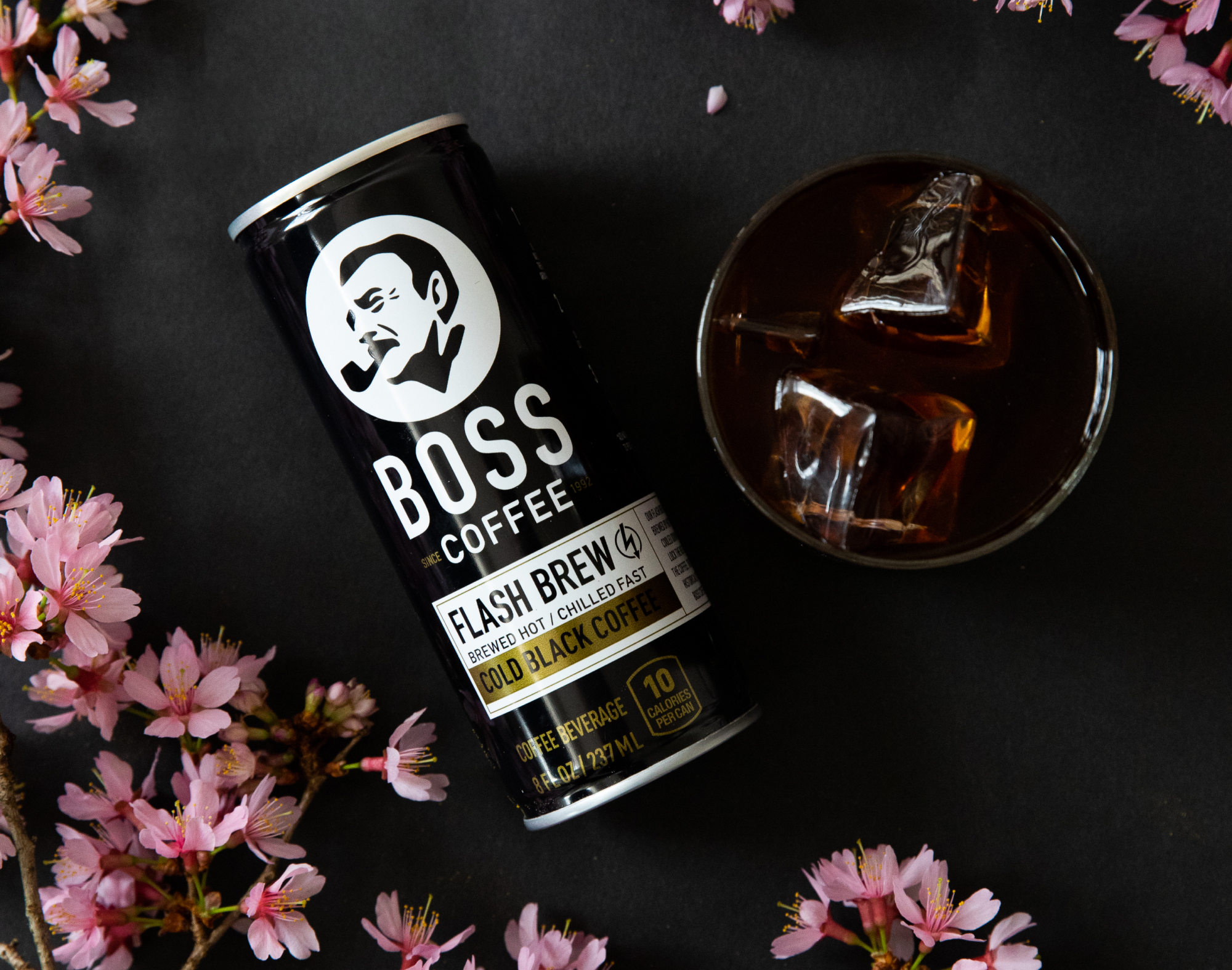 A moody top-down shot of a BOSS Coffee can and a rocks glass of coffee on a black background surrounded by pink flowers