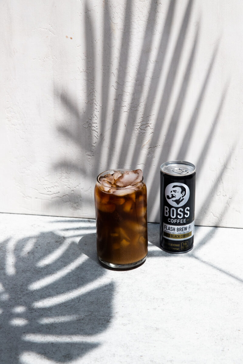 BOSS Coffee can next to a glass of iced coffee on a concrete scenery with shadows from tropical foliage.