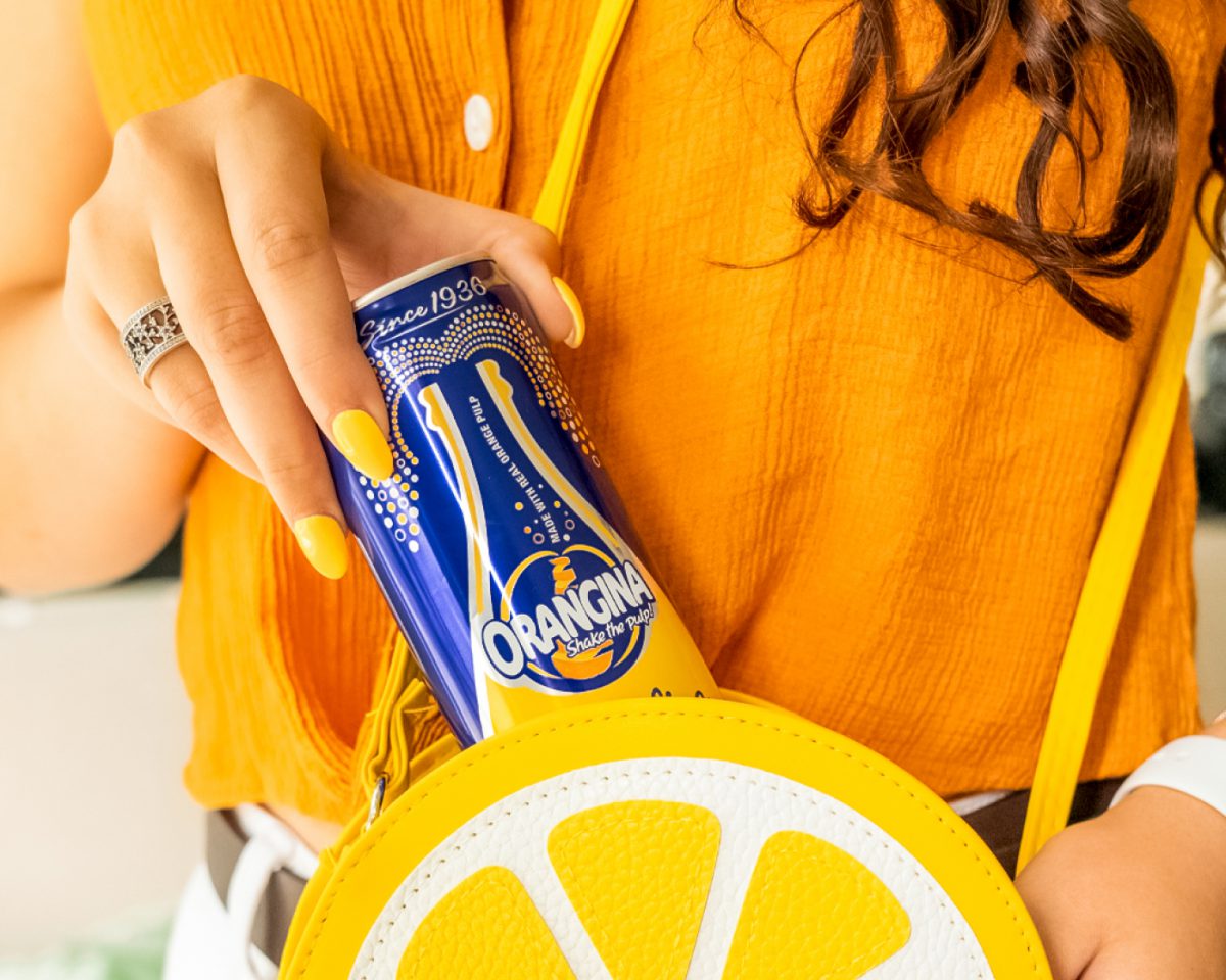 A woman with yellow polished nails, wearing a light orange blouse is pulling an Orangina canned beverage out of her lemon purse