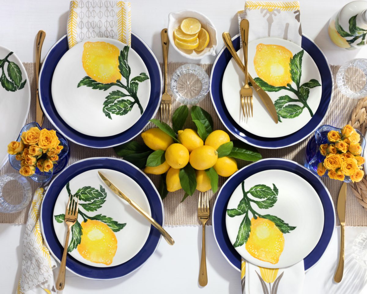 Vietri's lemon print plateware and gold utensils, styled on a beautiful top-down table setting with yellow flowers and lemons.
