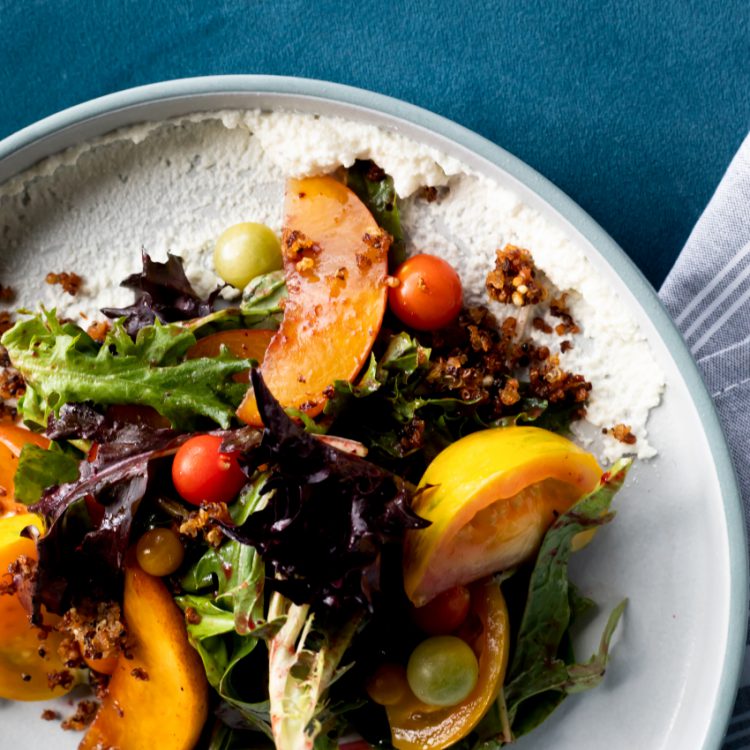 A highly styled top-down photo of a grilled peach salad with tomatoes, arugula, on a white ceramic plate and a rich blue tablecloth.