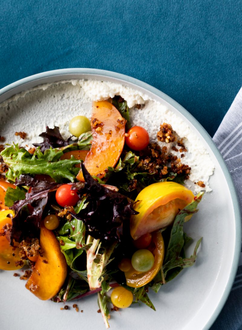 A highly styled top-down photo of a grilled peach salad with tomatoes, arugula, on a white ceramic plate and a rich blue tablecloth.