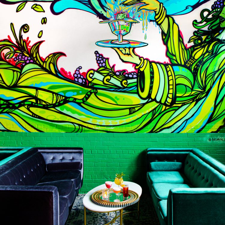 A vibrant, abstract mural in the focus of the restaurant setting with two velvet couches and a tray of assorted cocktails on a coffee table.