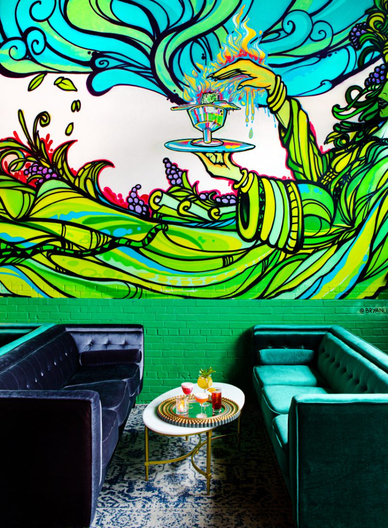 A vibrant, abstract mural in the focus of the restaurant setting with two velvet couches and a tray of assorted cocktails on a coffee table.