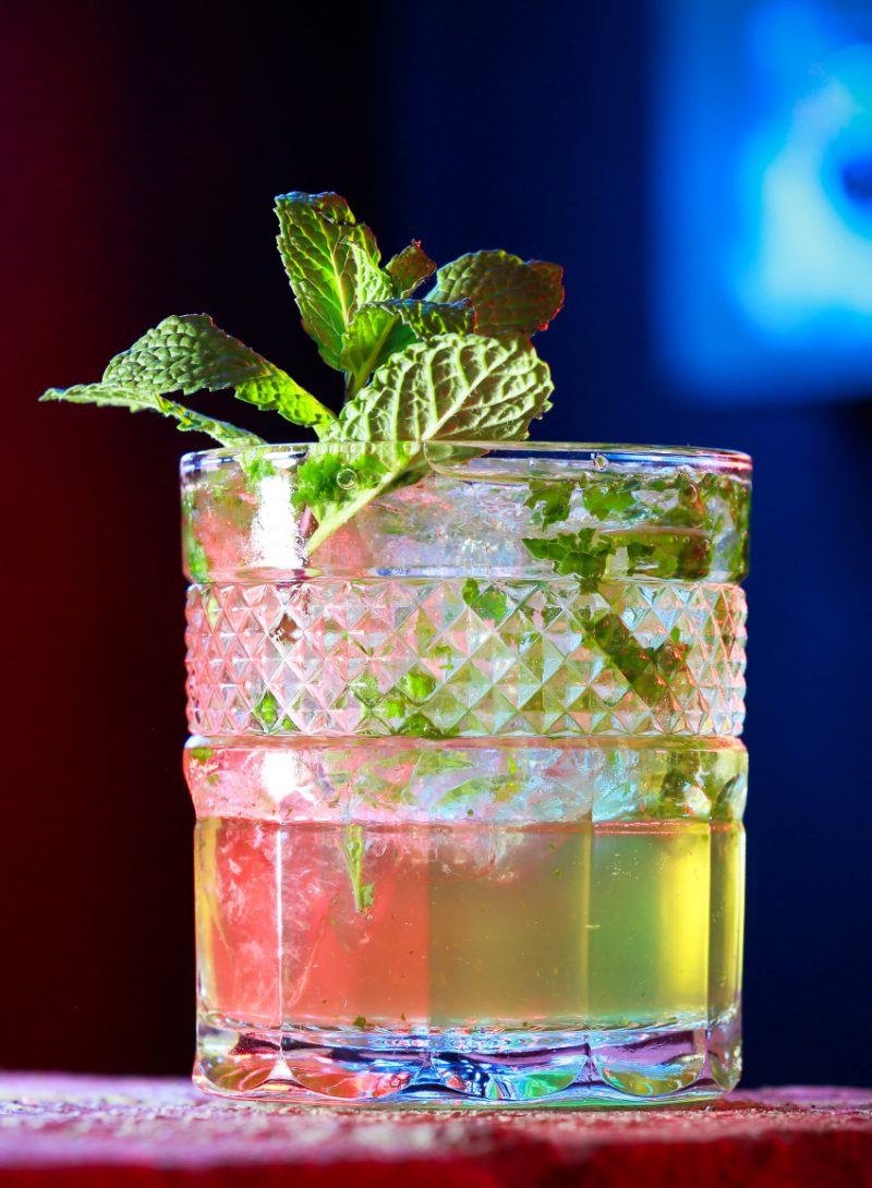 A cocktail in a rocks glass, eye-level with the camera in a dark atmosphere with mint leaves as garnish.