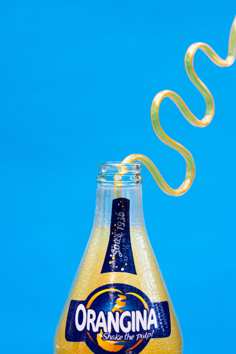 Close up of a bendy straw coming out of an Orangina bottle on a bright blue background