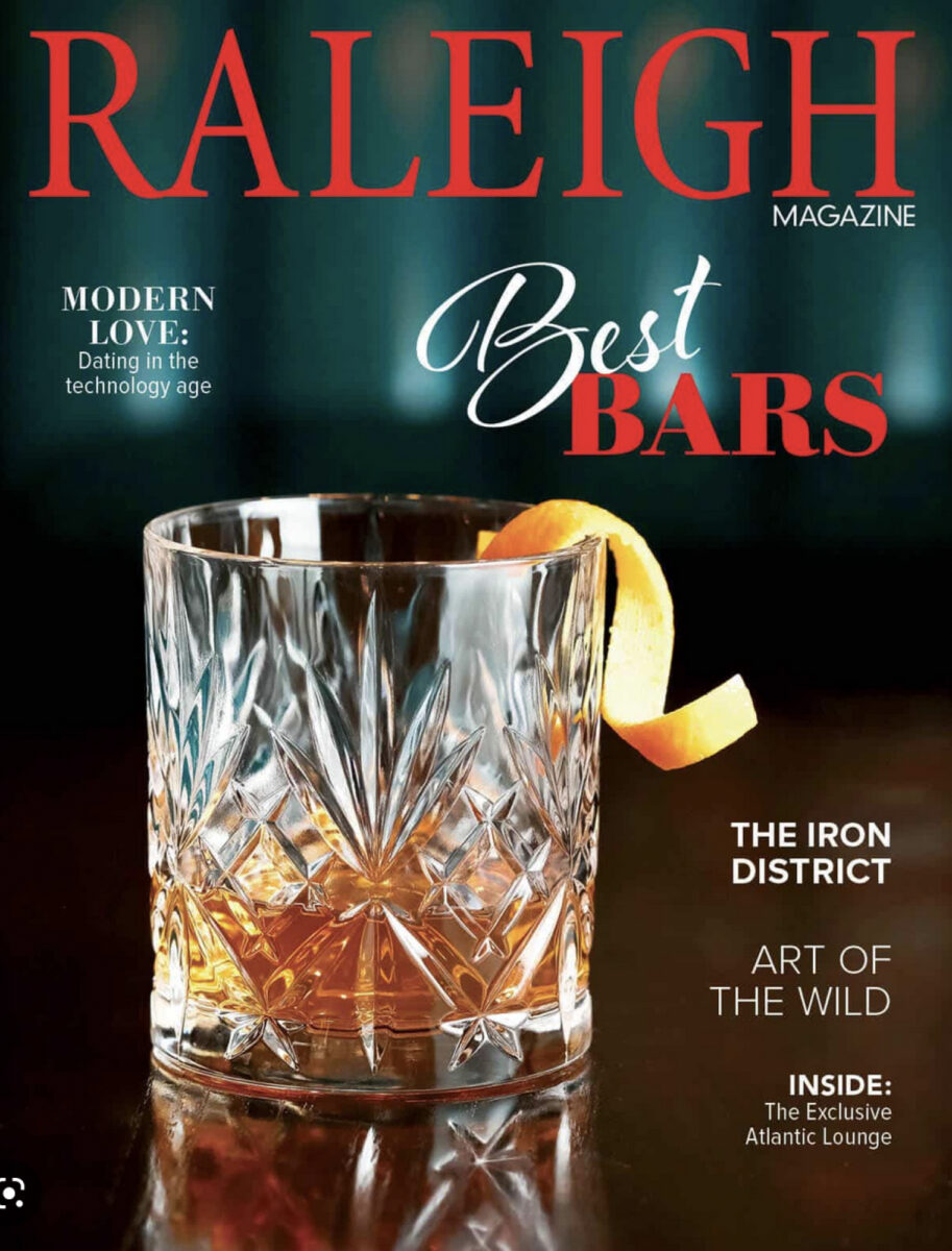 A rocks glass with a shot of whiskey in a dark setting with a headline of Best Bars – Raleigh Magazine