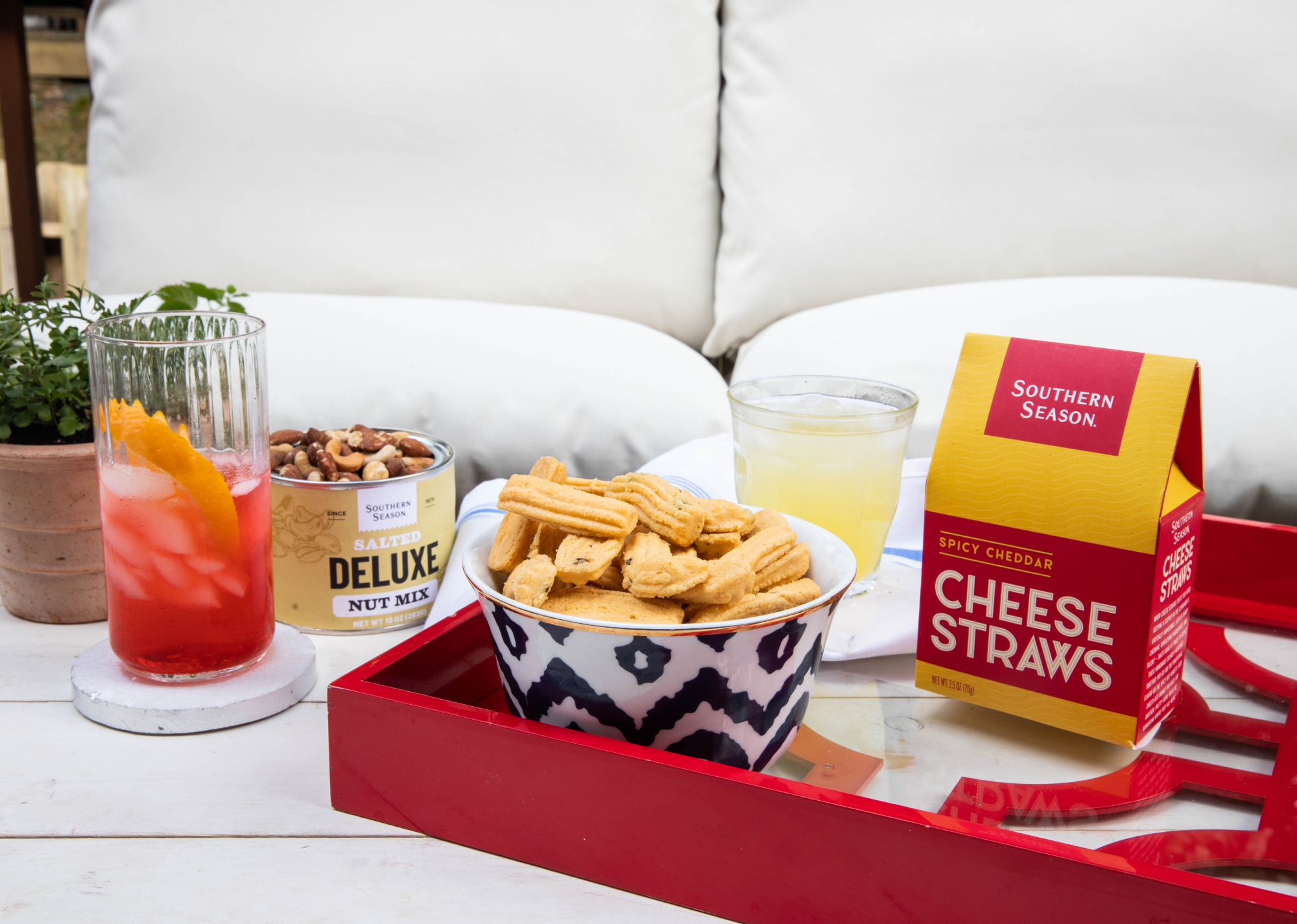 Southern Season's Deluxe Salted Nut Mix and Cheese Straws set out on a red tray on a patio table with lemonade