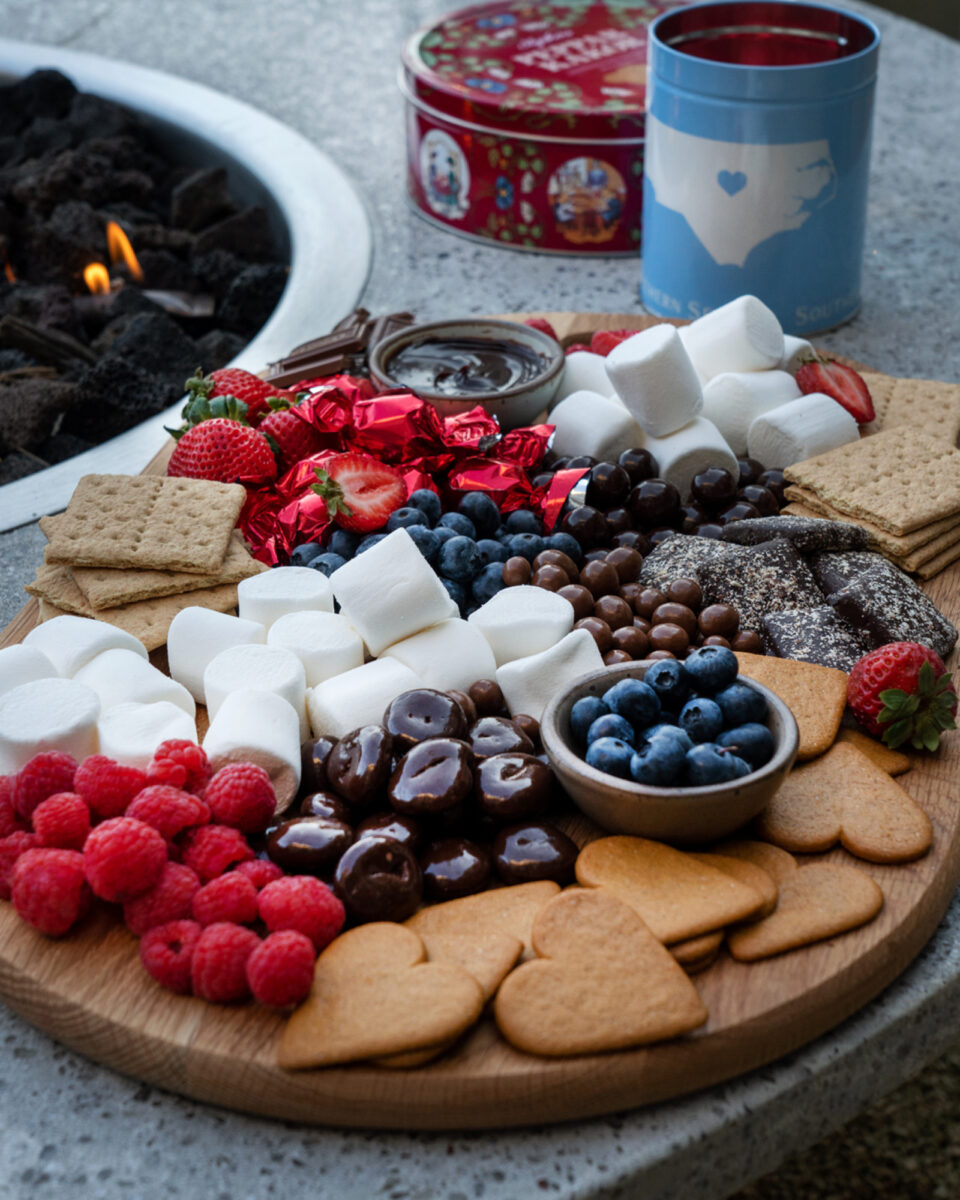 A fancy smore's board with marshmallows, fresh fruit, heart shaped cookies, graham crackers, chocolate covered nuts and more, next to a smouldering campfire.