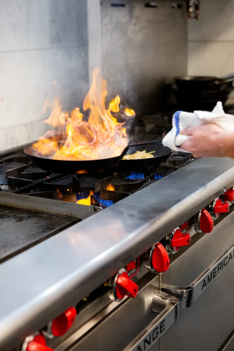 A close up photo of a cook holding a frying pan that is engulfed in flames over an American Range cooktop