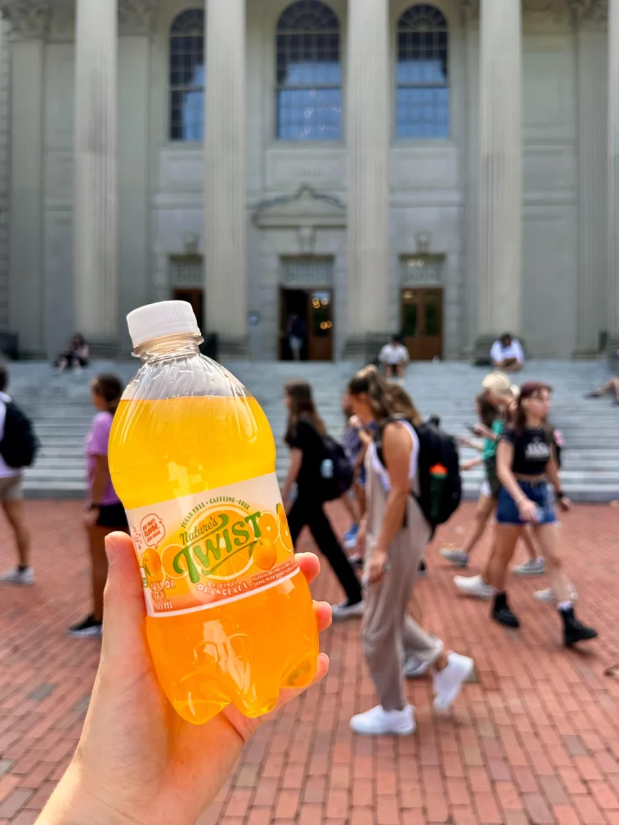 A hand holding a bottle of Nature's Twist Orangeade in front of the busy steps of a university